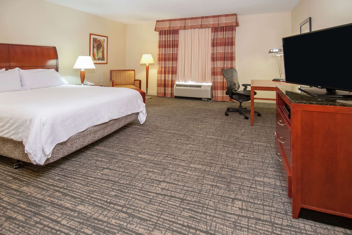 A hotel room with a large bed, a desk with a television, a chair, a lamp, and an air conditioning unit.