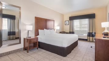 A hotel room with a large bed, bedside tables with lamps, a desk with a chair, a large mirror, and a window with curtains.