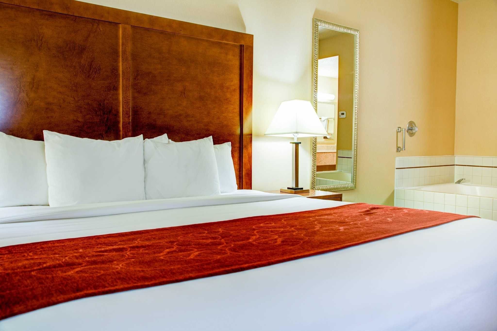 Habitación de hotel con una cama grande, cabecera de madera, manta roja, lámpara de noche, espejo y una bañera blanca.