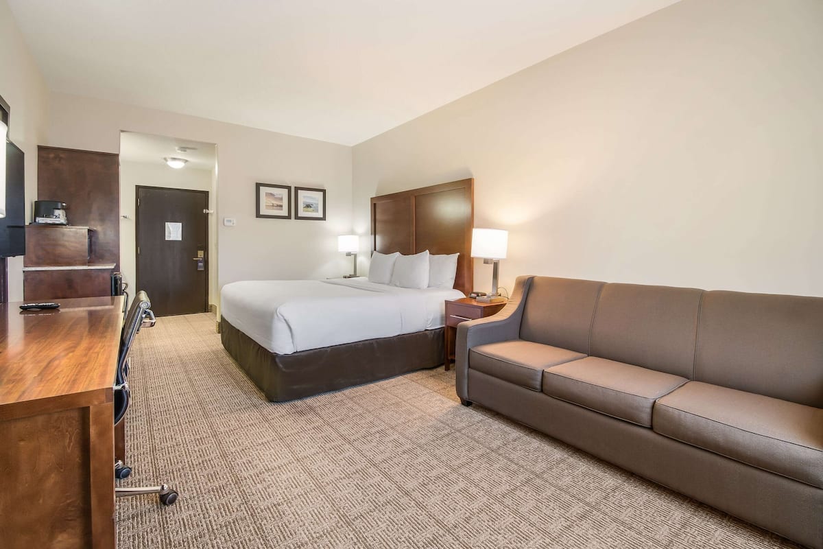 A hotel room with a bed, a brown leather sofa, a desk with a chair, and a small table with a lamp.
