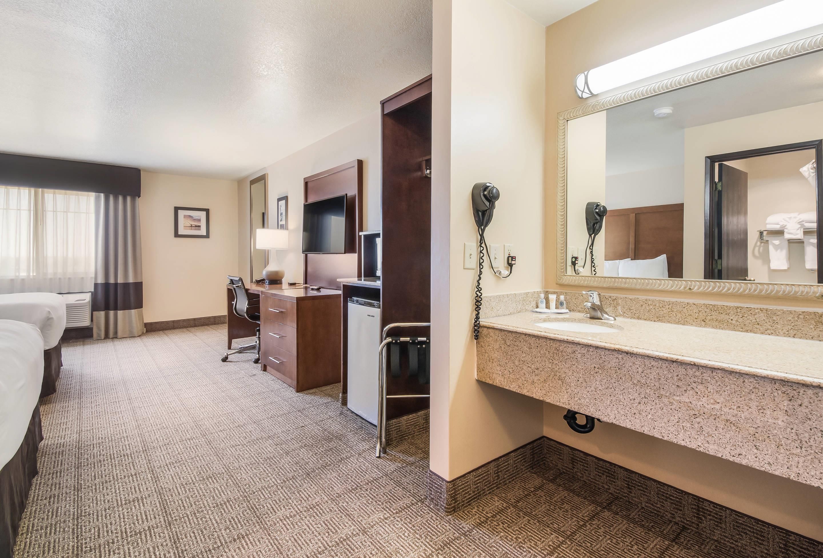 A hotel room with a bed, desk, chair, and a bathroom with a sink and mirror.