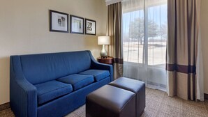 Una habitación con un sofá azul, un pequeño reposapiés, una lámpara y una ventana con cortinas.