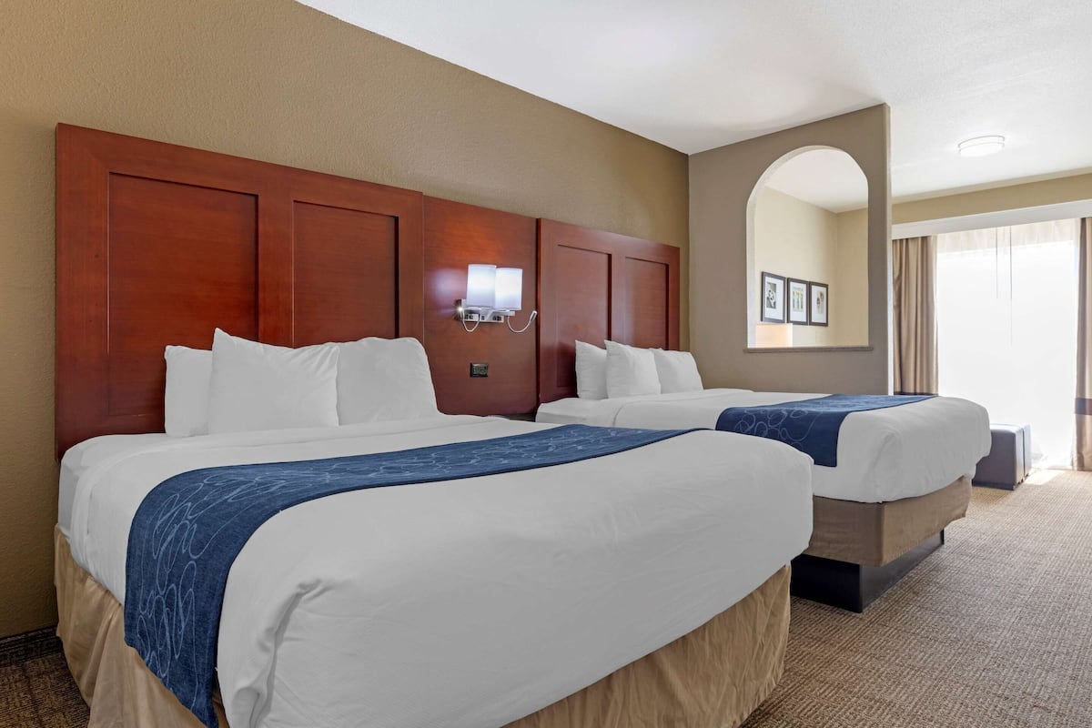 A hotel room with two beds, a wooden headboard, a wall-mounted light, and a large window with curtains.