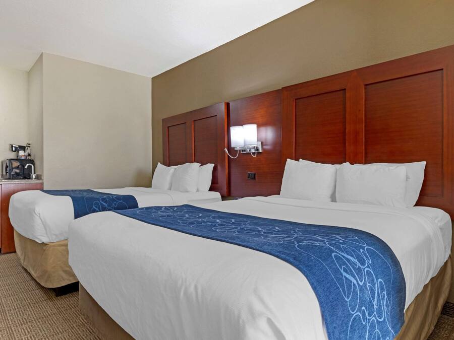 A hotel room with two beds, a wooden headboard, a bedside table with a lamp, and a small desk with a coffee maker.