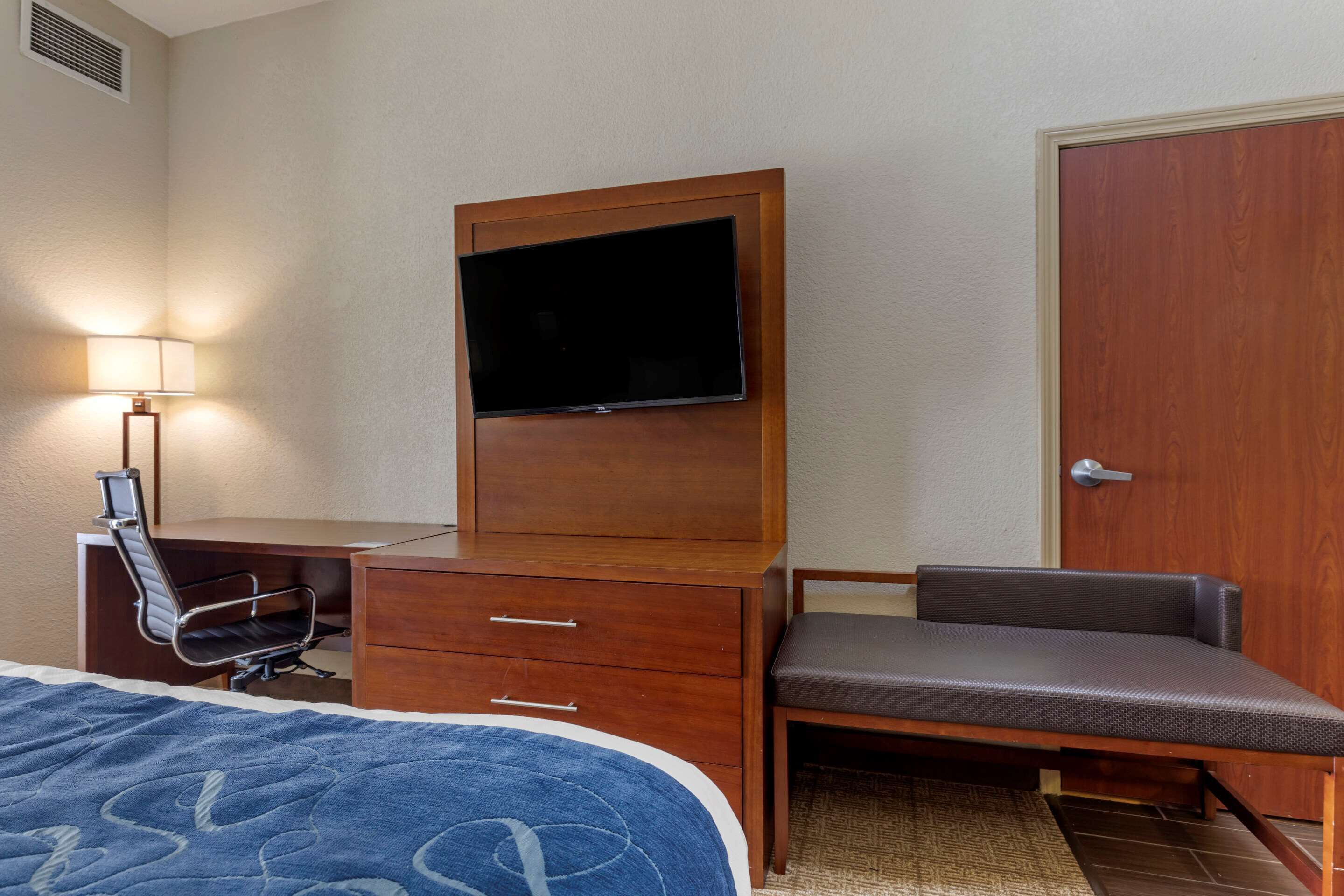 A hotel room with a bed, a desk with a chair, a television mounted on a wooden cabinet, and a door.