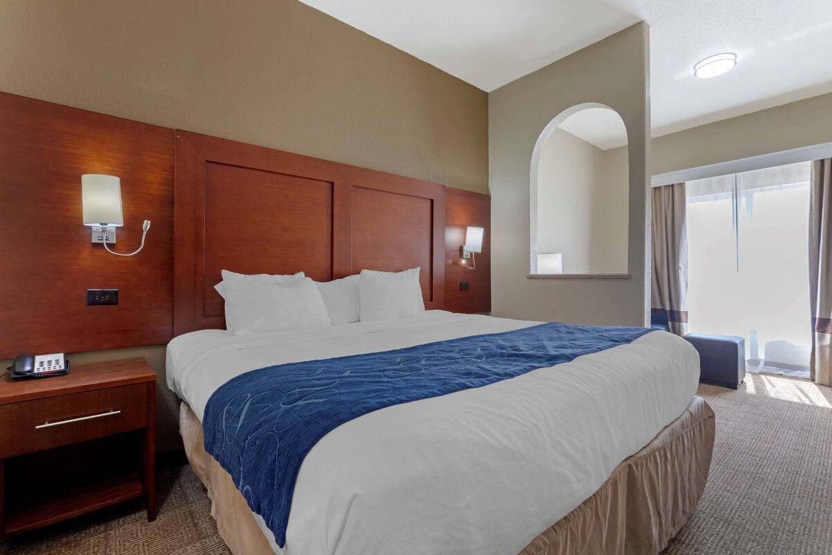 A hotel room with a large bed, wooden headboard, bedside lamps, a telephone, and a view of the outside through a large window.