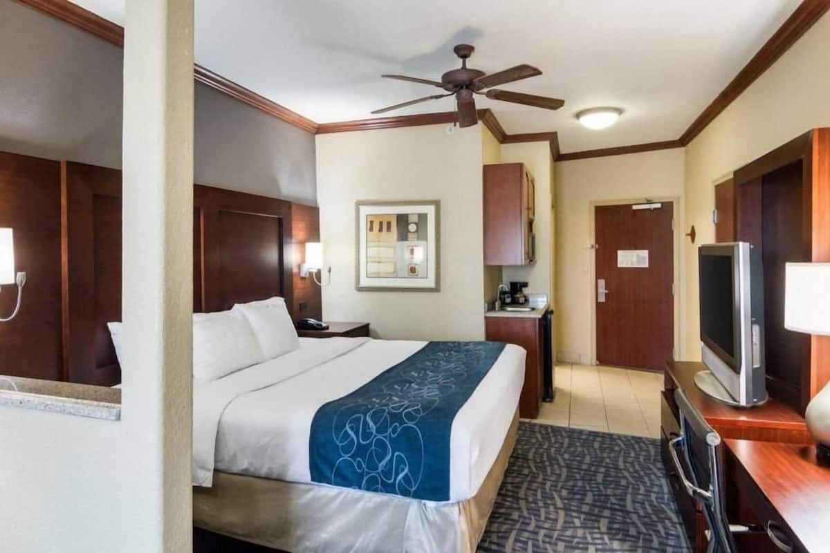A hotel room with a bed, a desk, a television, a ceiling fan, and a small kitchenette.