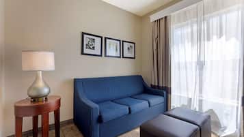 Ein Hotelzimmer mit einem blauen Sofa, einem kleinen Holztisch mit Lampe, einem schwarzen Hocker und einem großen Fenster mit durchsichtigen Vorhängen.