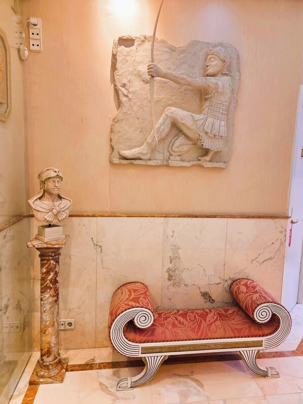 Una habitación con piso de mármol, un banco tapizado en rojo, un pedestal de mármol con un busto y un relieve escultórico en la pared.