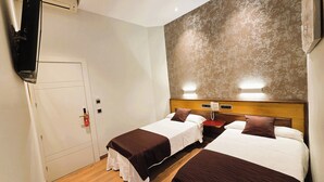 A hotel room with two beds, a wooden floor, a wall-mounted TV, and a door with a fire alarm.