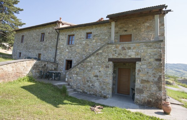Un edificio de piedra con puerta de madera, una escalera y un patio con mobiliario exterior.