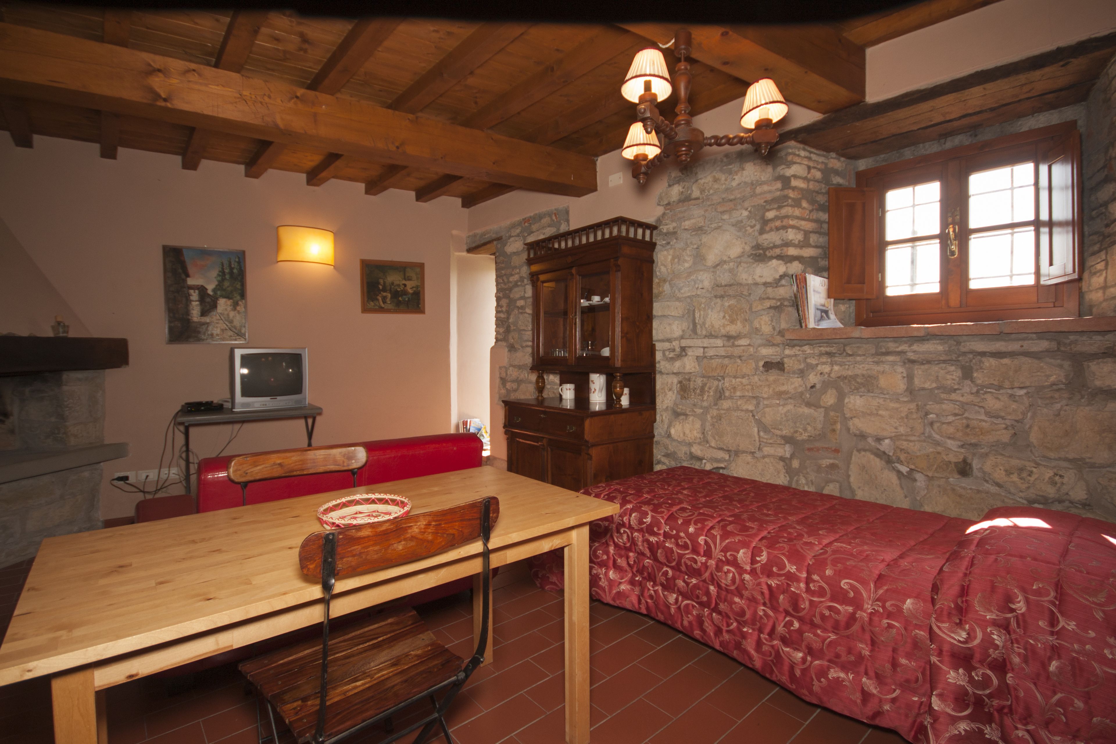 A cozy room with a wooden table, a red sofa, a stone wall, and a window with wooden shutters.