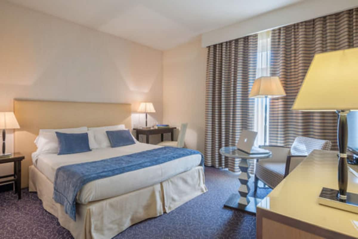 Habitación de hotel con cama, dos mesitas de noche con lámparas, un escritorio con silla y una ventana con cortinas.