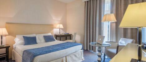 Quarto de hotel com uma cama, duas mesas de cabeceira com luminárias, uma escrivaninha com cadeira e uma janela com cortinas.