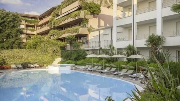 Een hotel met een zwembad, omringd door ligbedden en parasols, met groen en een meer verdiepingen tellend gebouw op de achtergrond.