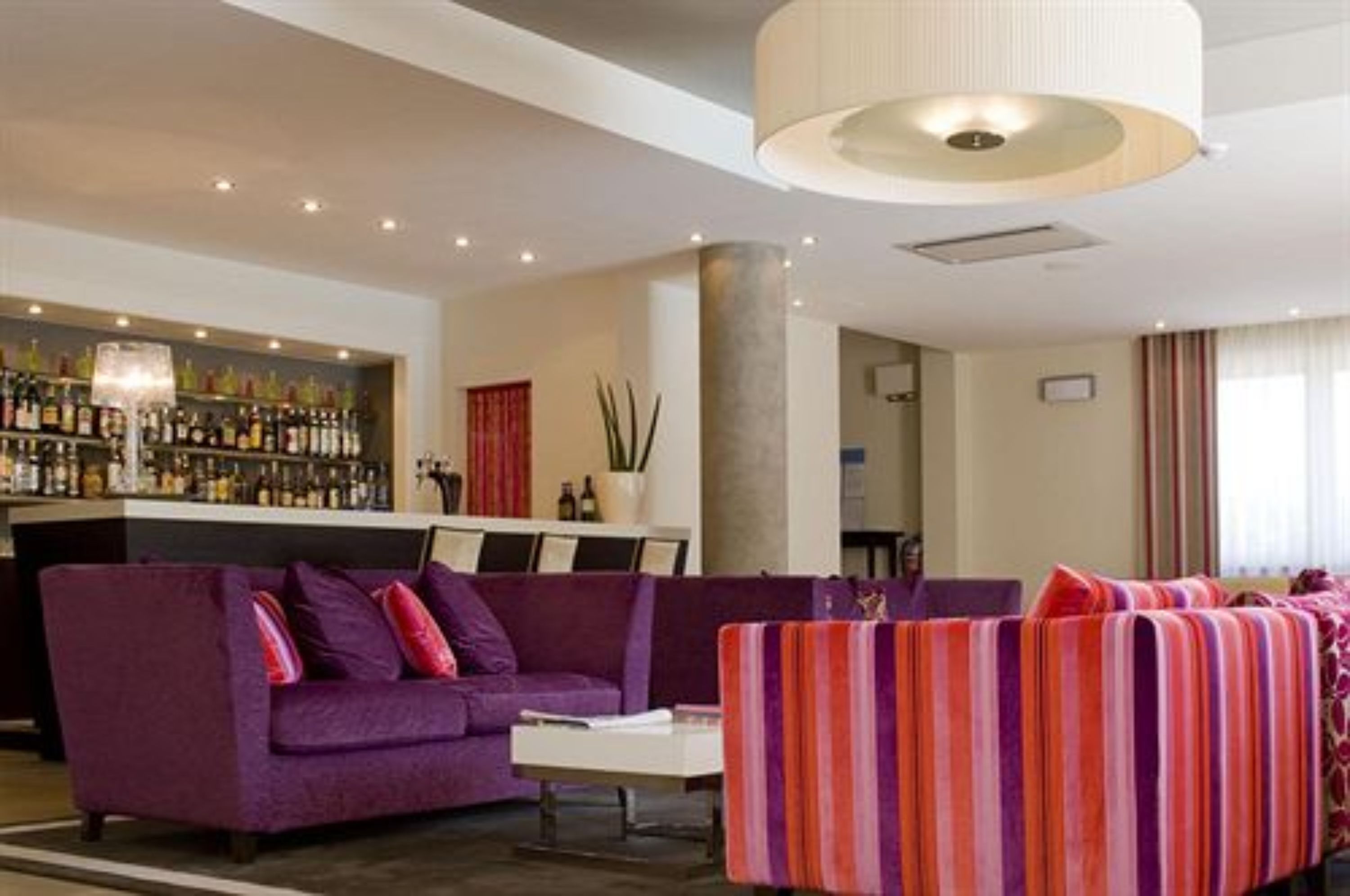Un vestíbulo de hotel con un sofá morado, un sillón a rayas, una zona de bar con varias botellas y un pilar grande.