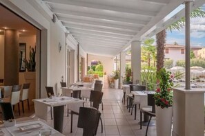 Un espacio de comedor al aire libre con techado, mesas y sillas, rodeado de plantas y vistas al jardín.