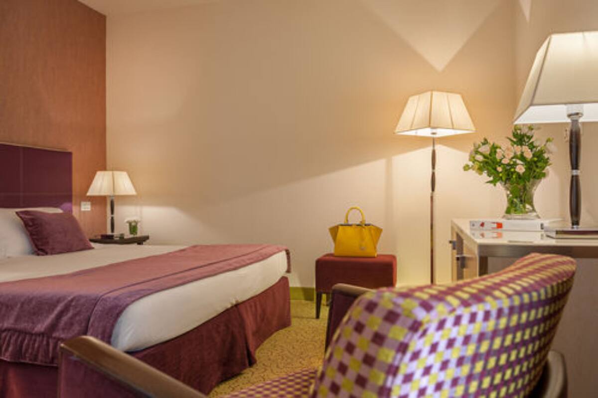 Habitación de hotel con cama, mesita de noche, lámpara, silla y escritorio con lámpara y flores.