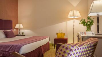 A hotel room with a bed, bedside table, lamp, chair, and a desk with a lamp and flowers.