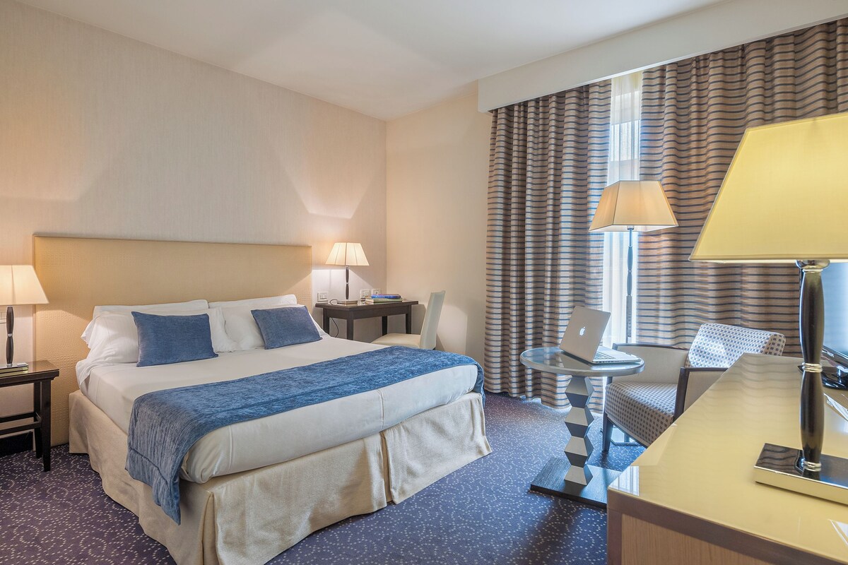 Habitación de hotel con una cama grande, dos mesitas de noche con lámparas, un escritorio con silla y una ventana con cortinas.