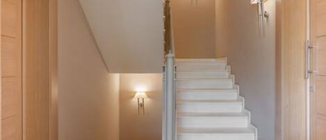 A staircase with white steps and a wooden handrail, flanked by light-colored walls and two wall-mounted light fixtures.