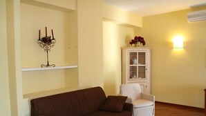 Een woonkamer met een bruine bank, een witte fauteuil, een witte kast met glazen deuren, een kandelaar met kaarsen en een wandlamp.