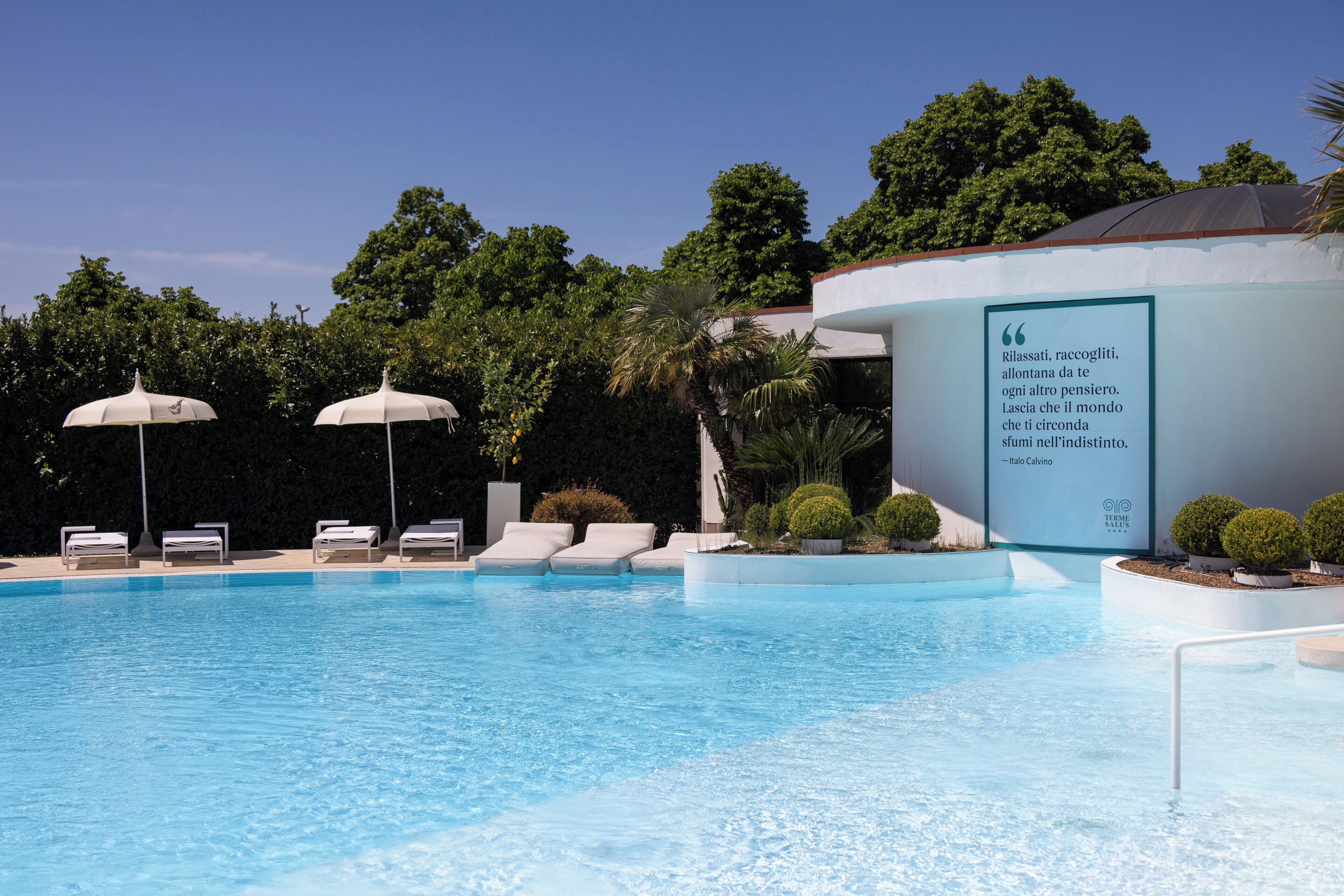 Piscina con lettini e ombrelloni, immersa nel verde e con una citazione su un muro.
