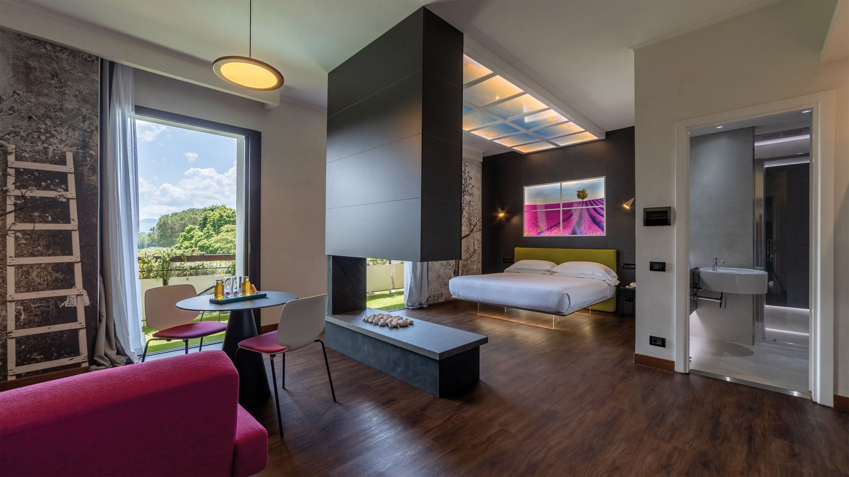 A modern hotel room with a bed, a small table, a chair, a large window with a view of greenery, and a skylight.