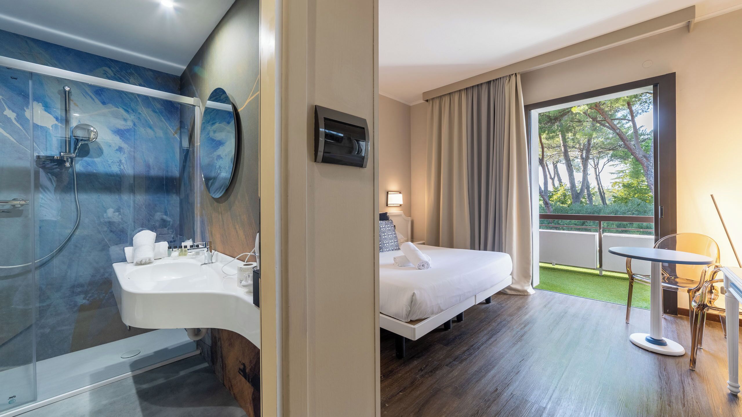 Habitación de hotel con baño, cama, una mesita y balcón con vista a árboles.
