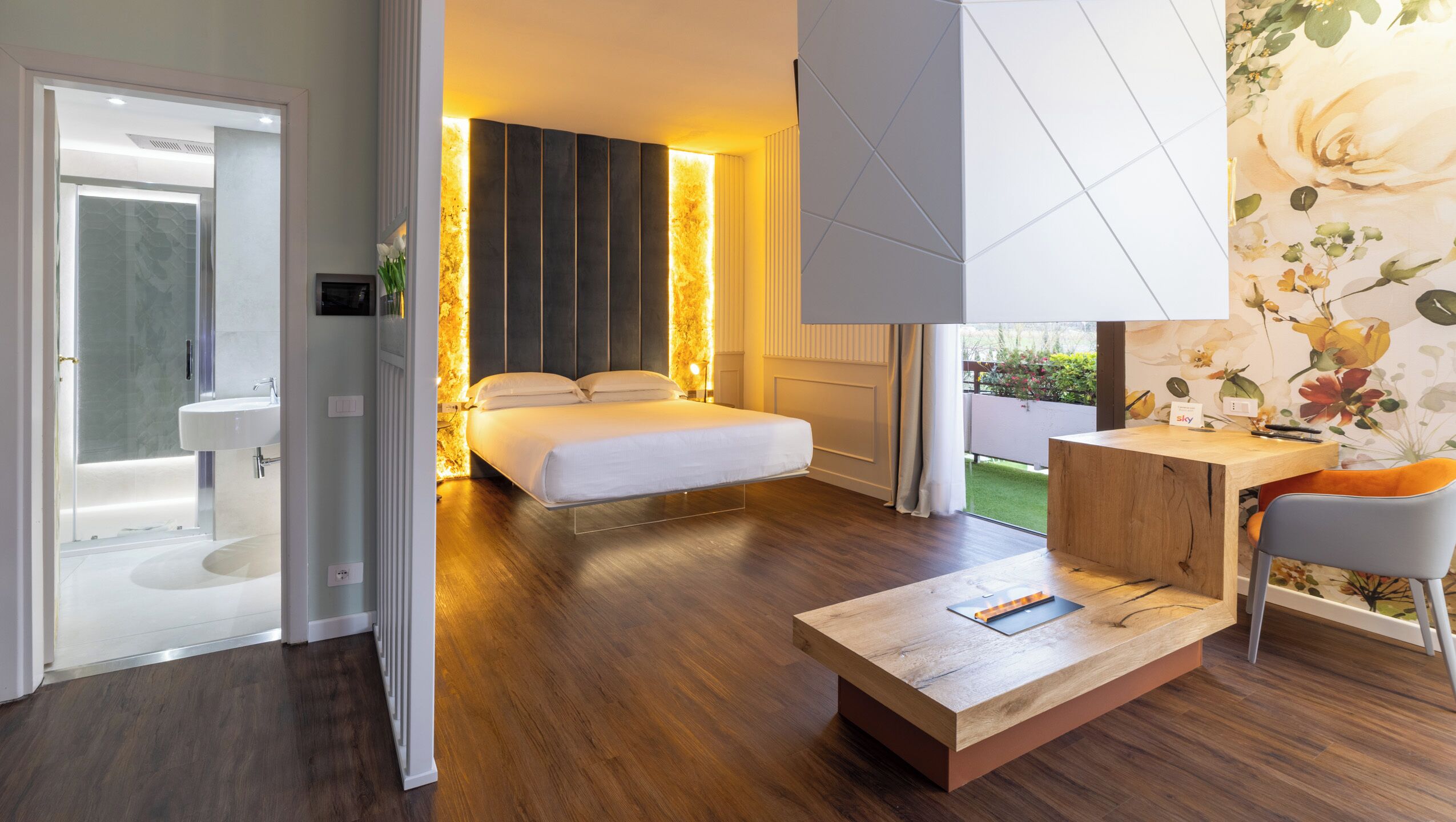 A modern hotel room with a wooden floor, a large bed, a desk with a chair, and a view of a garden.