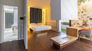 A modern hotel room with a wooden floor, a large bed, a desk with a chair, and a view of a garden.