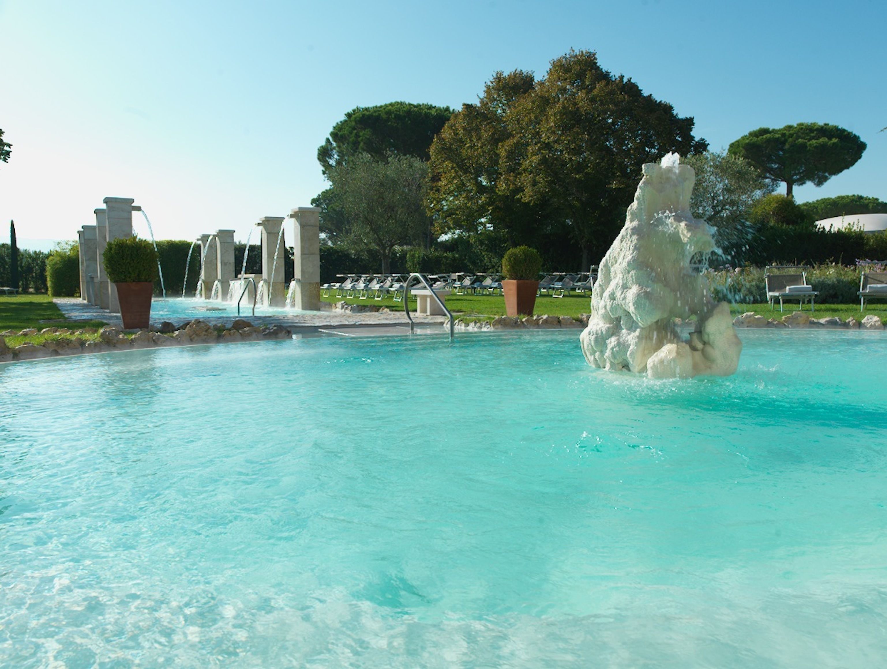 A pool with a central water feature and surrounding lounge chairs.
