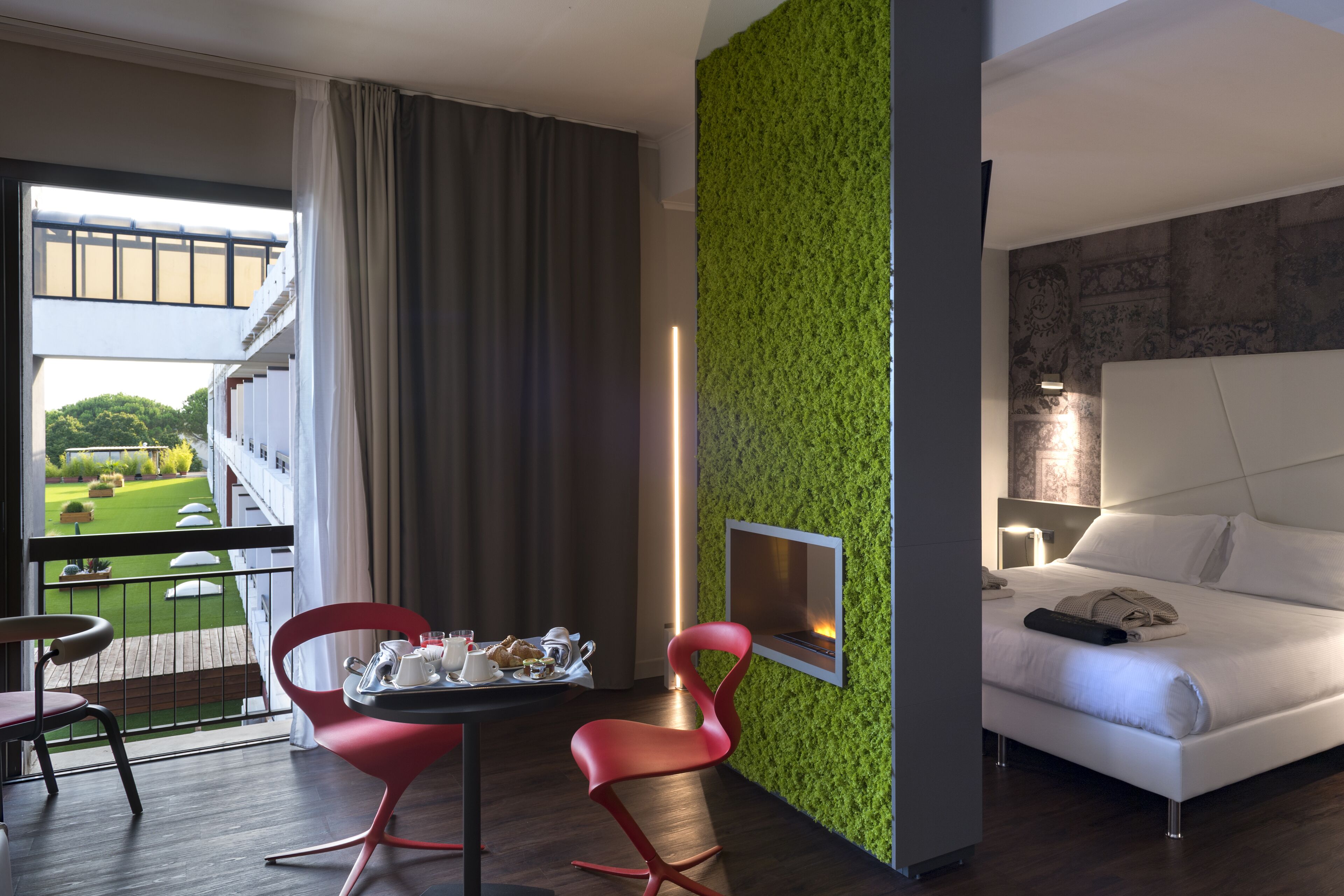 A hotel room with a balcony, a dining area with red chairs, a bed with white linens, and a green textured wall.