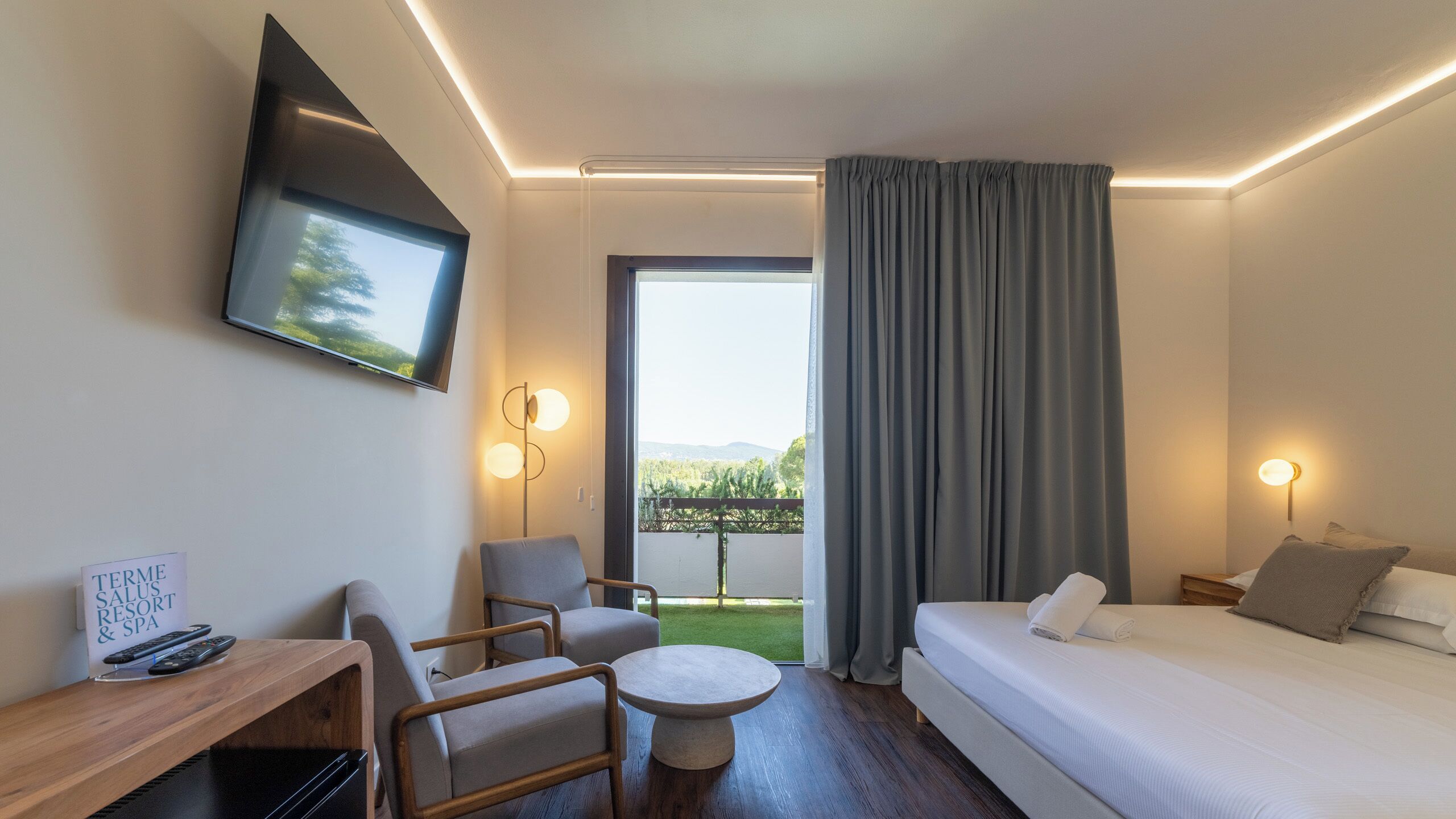 A hotel room with a bed, a desk with a TV, a chair, a small table, and a view of a green landscape through the window.