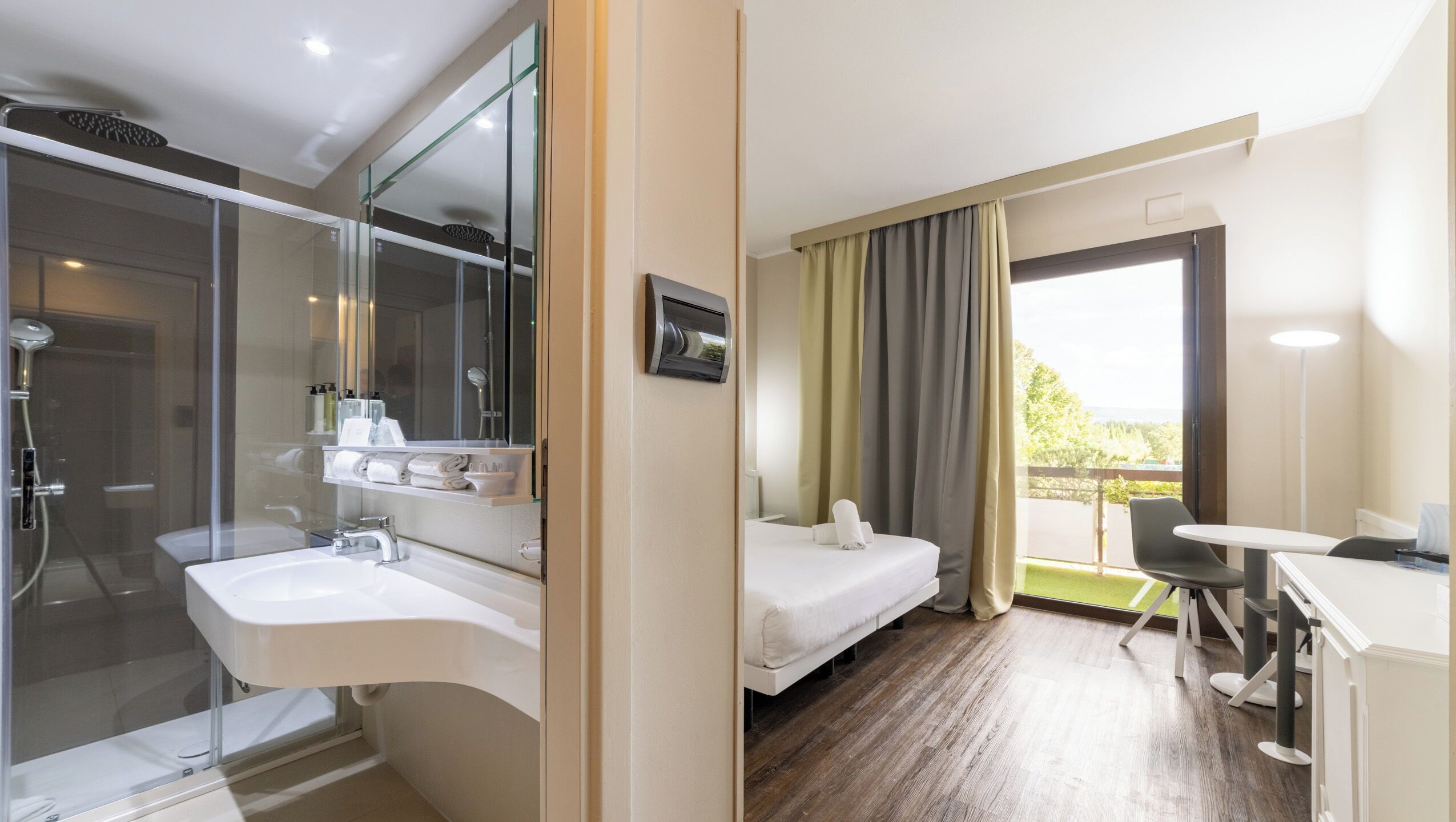 Hotelkamer met douchecabine van glas, een wastafel, een bed, een bureau en uitzicht op de omgeving.
