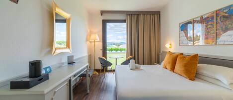 A hotel room with a bed, a desk, a chair, a lamp, and a view of the outdoors.