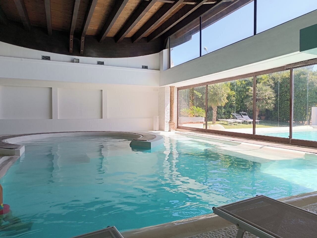 Una piscina interior con agua azul cristalina, ventanas amplias y techo de madera.