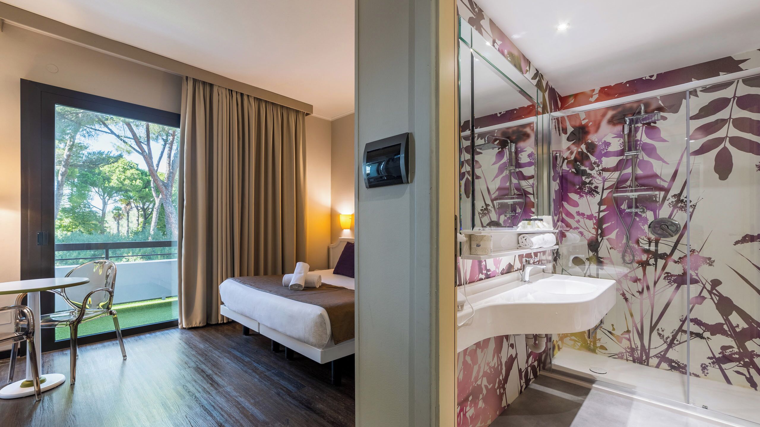A hotel room with a bed, a chair, a table, a window with curtains, a bathroom with a sink and shower, and a mirror.