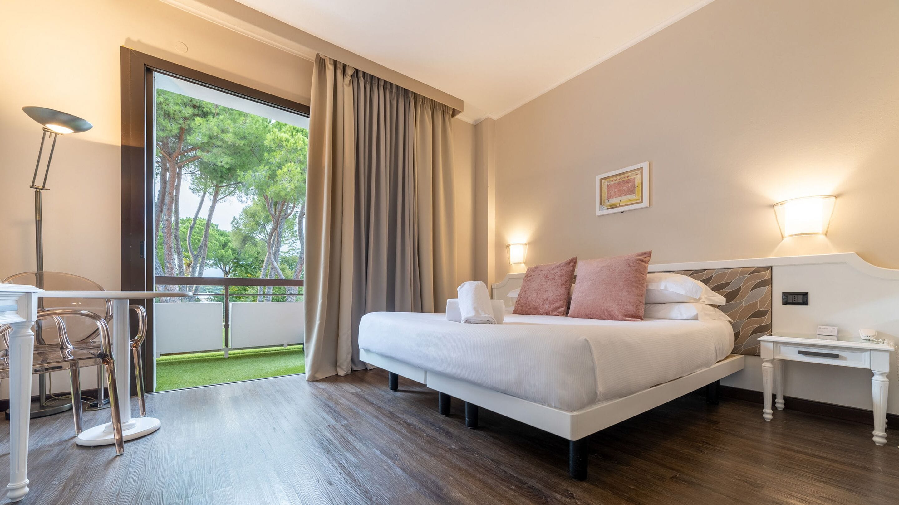 Habitación de hotel con cama, mesita de noche, lámpara y vistas al exterior.