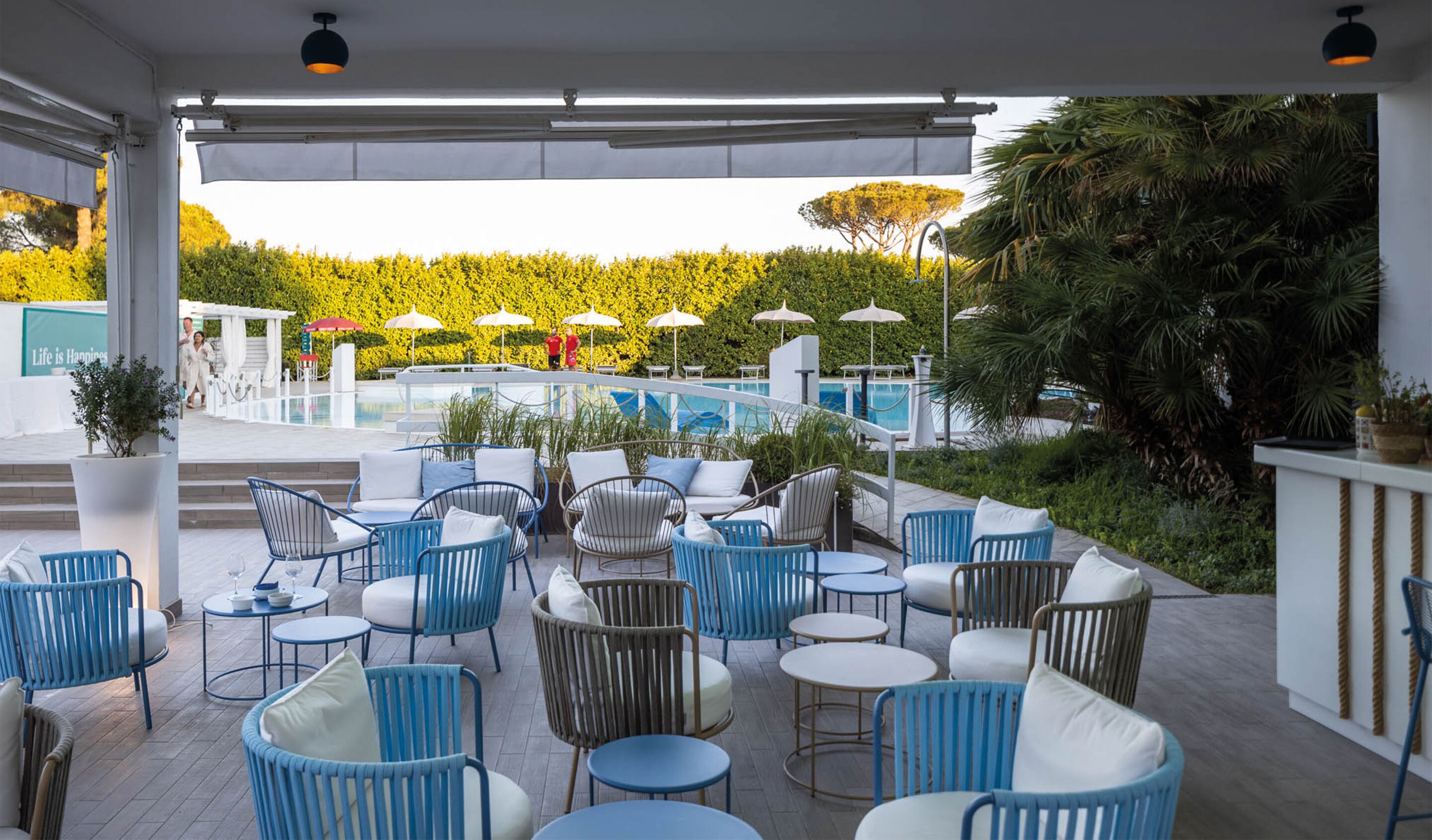 Ein Außenbereich mit blauen und weißen Stühlen und Tischen, mit Blick auf einen Pool und umgeben von viel Grün.
