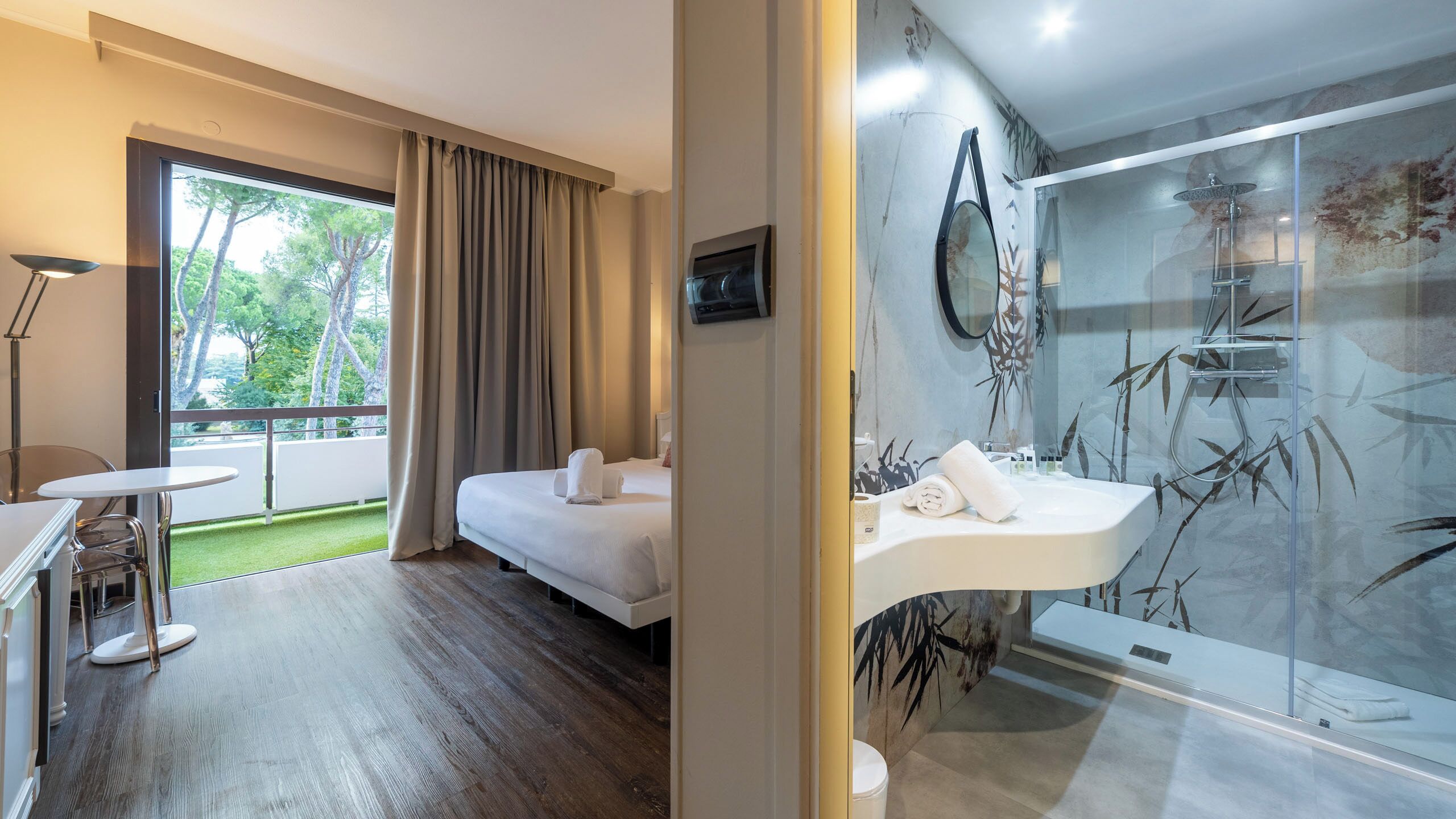 Ein Hotelzimmer mit Bett, kleinem Tisch, Stuhl, einem Balkon mit Vorhängen sowie einem Badezimmer mit Waschbecken und Dusche.