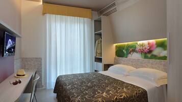 A hotel room with a bed, a desk with a hat and sunglasses, a TV displaying a cityscape, and a wall-mounted picture of a bee on a flower.