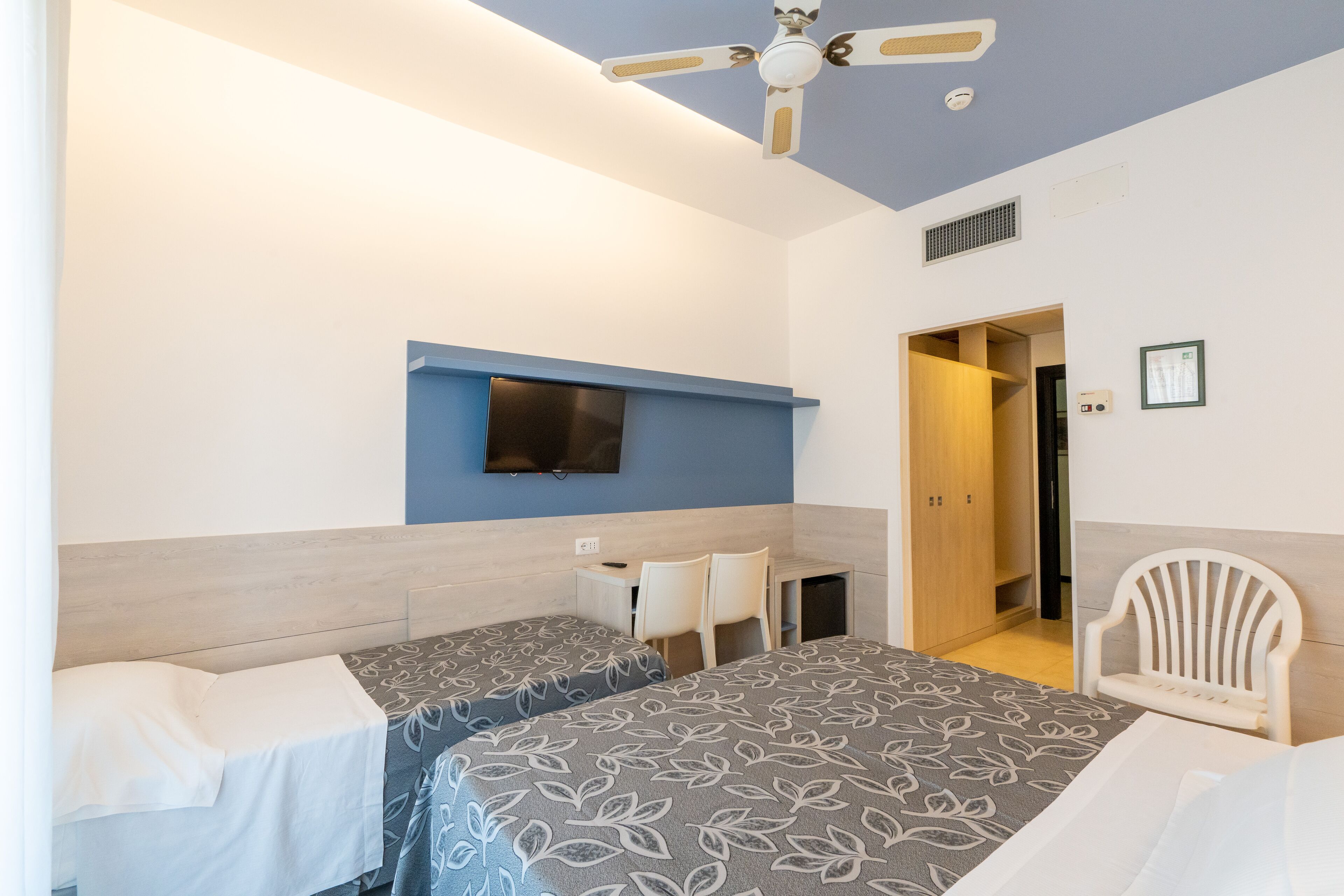 A hotel room with two beds, a desk, a chair, a TV, and a ceiling fan.
