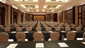Un salón de conferencias con filas de mesas y sillas, equipado con botellas de agua y documentos, con decoración tradicional y una pantalla grande al frente.