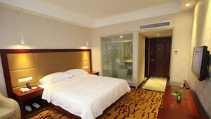 Habitación de hotel con una cama grande, una mesita de noche con teléfono y lámpara, una zona de estar con mesa y sillón, y un televisor en la pared.