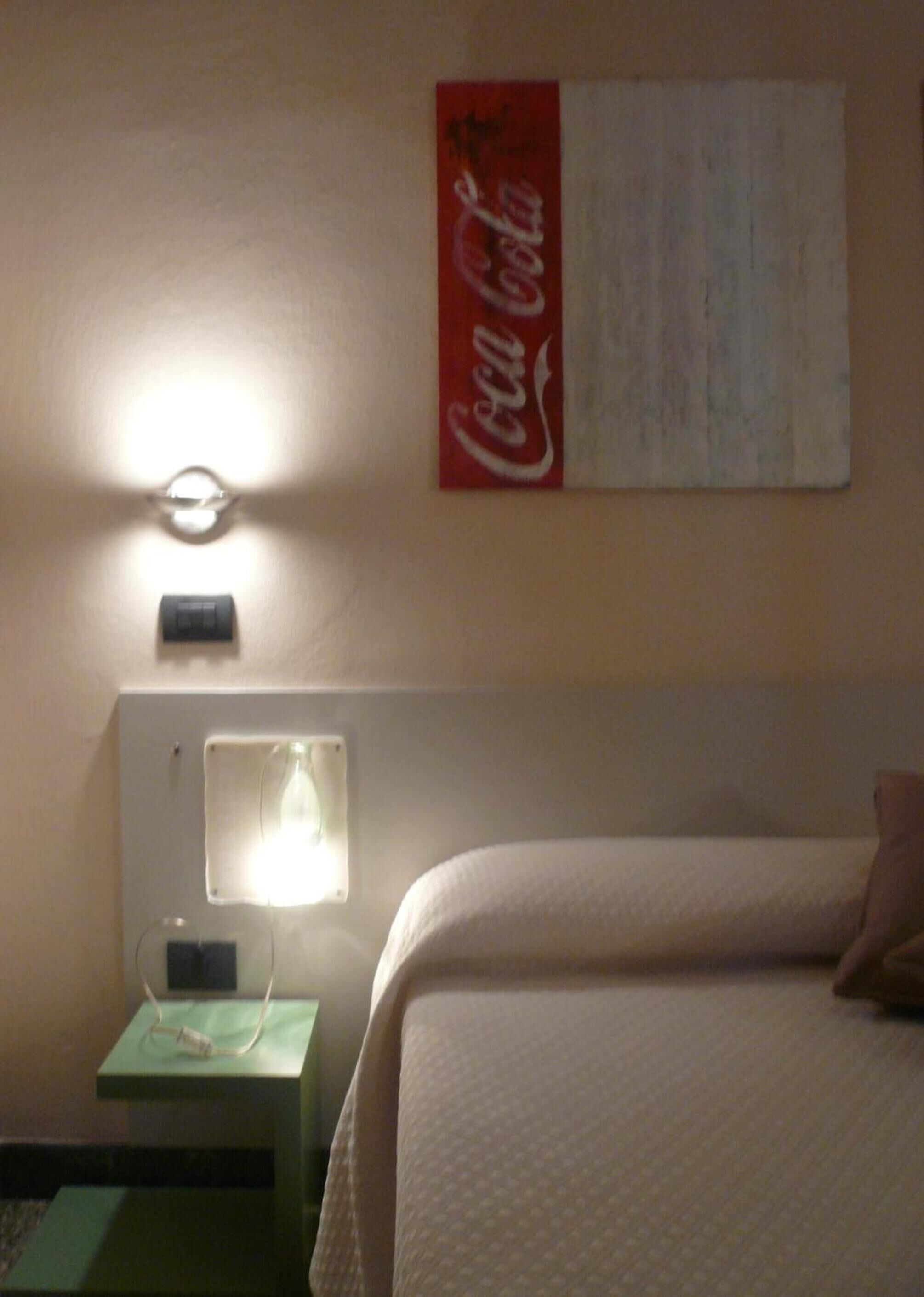 A hotel room with a bed, a nightstand, a wall-mounted lamp, and a Coca-Cola sign.