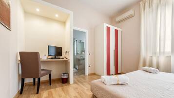Quarto de hotel com cama, escrivaninha com computador, cadeira e banheiro visível por uma porta aberta.