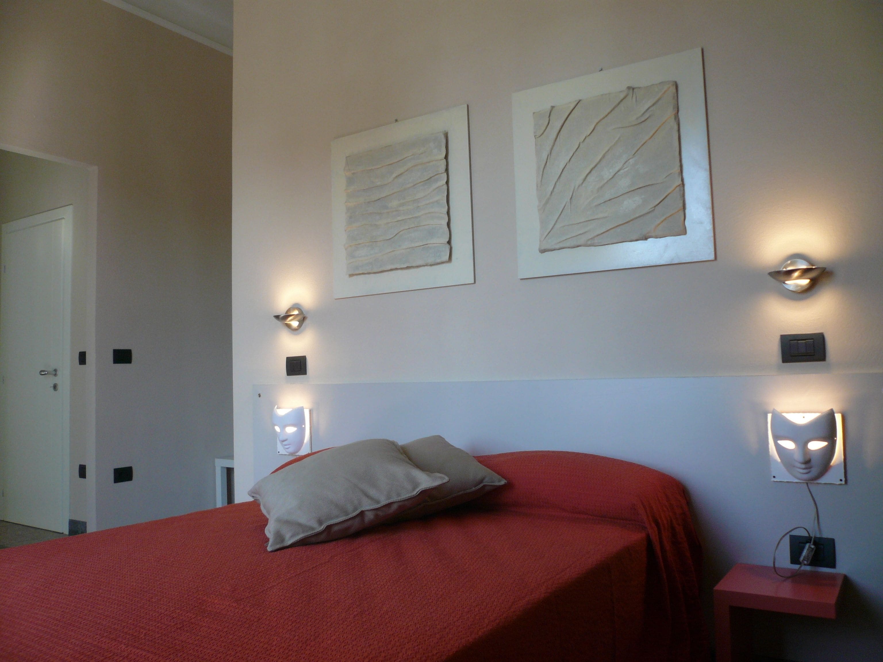 A bedroom with a red bedspread, two wall-mounted light fixtures, and two framed artworks.