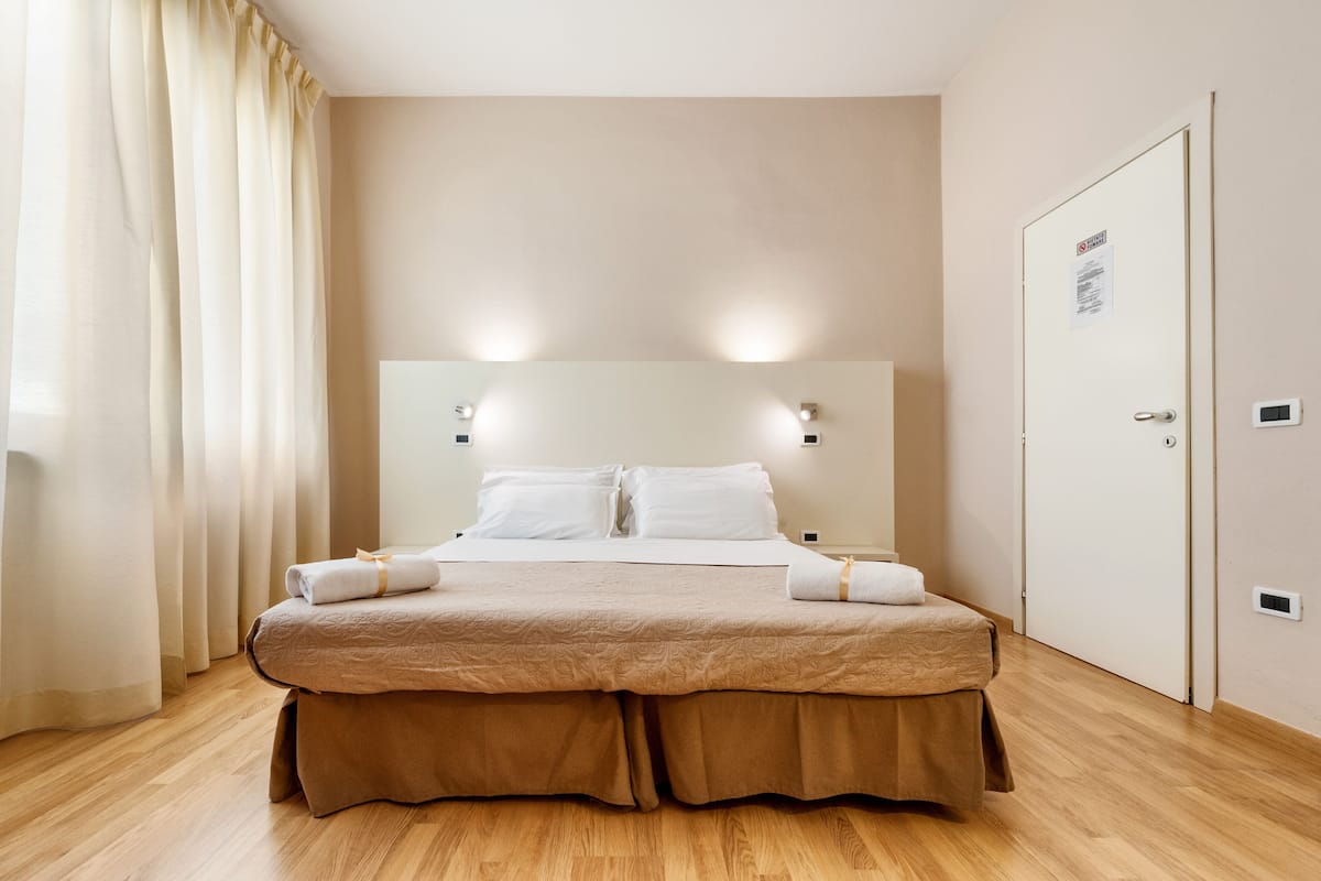 A double bed with a beige bedspread and two pillows, a bedside table with a lamp, and a door with a sign.