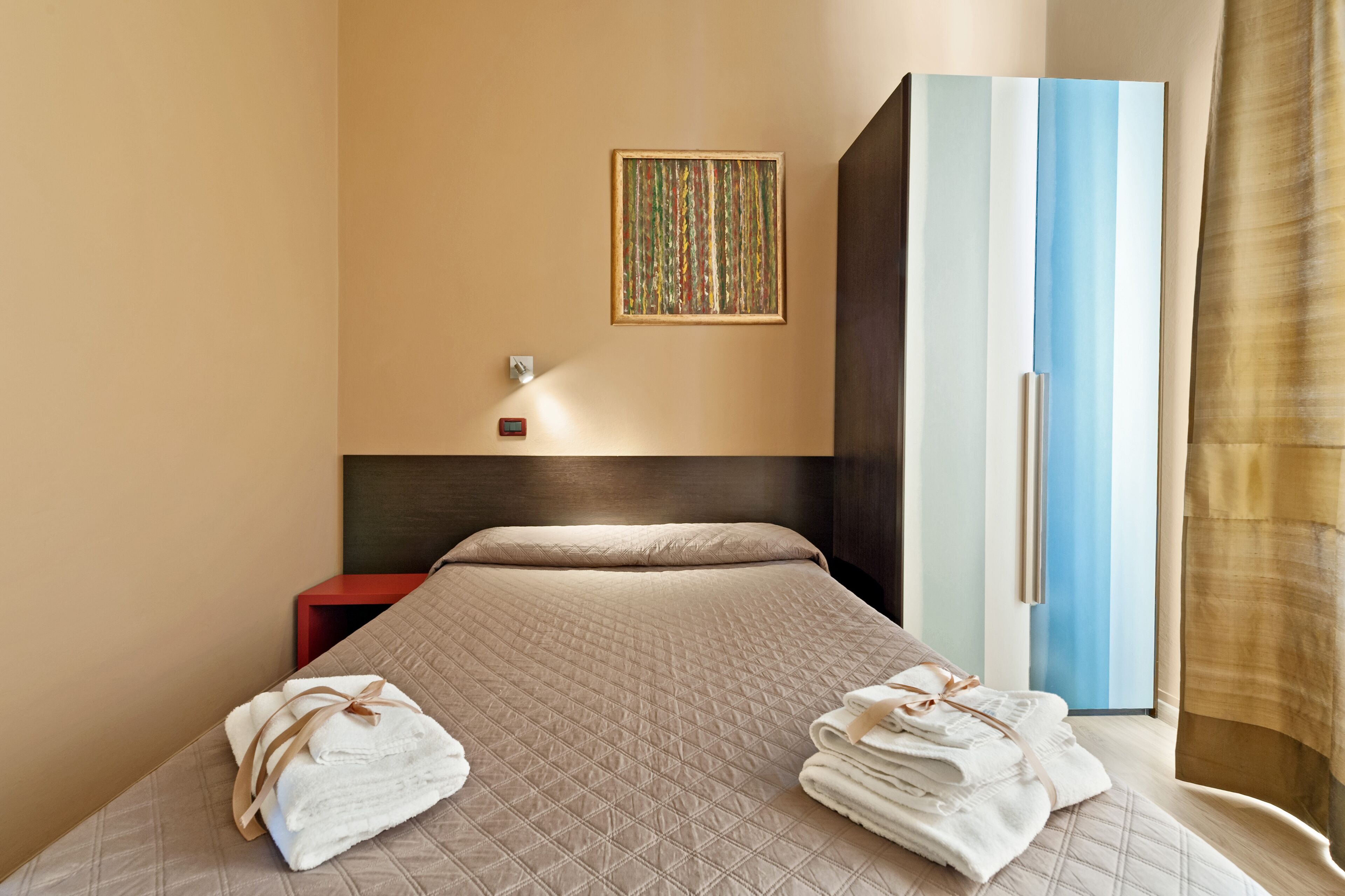 A hotel room with a bed, a wardrobe, a wall-mounted light, and a painting.